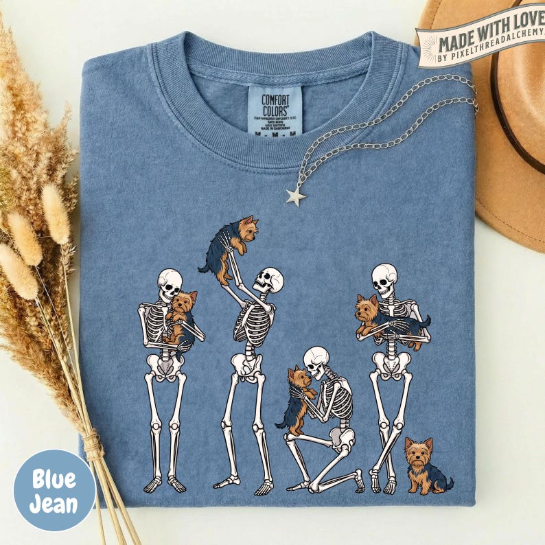 May include: A blue crew neck t-shirt featuring a graphic print of four white skeletons interacting with small brown Yorkshire Terrier dogs. One skeleton holds a dog overhead, another kneels to hold a dog, and two others hold dogs.