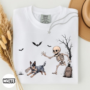 May include: A white t-shirt featuring a graphic print of a skeleton emerging from a grave near a bare tree, with a blue heeler dog running while holding a bone in its mouth. Two bats are flying in the background above the skeleton and dog.