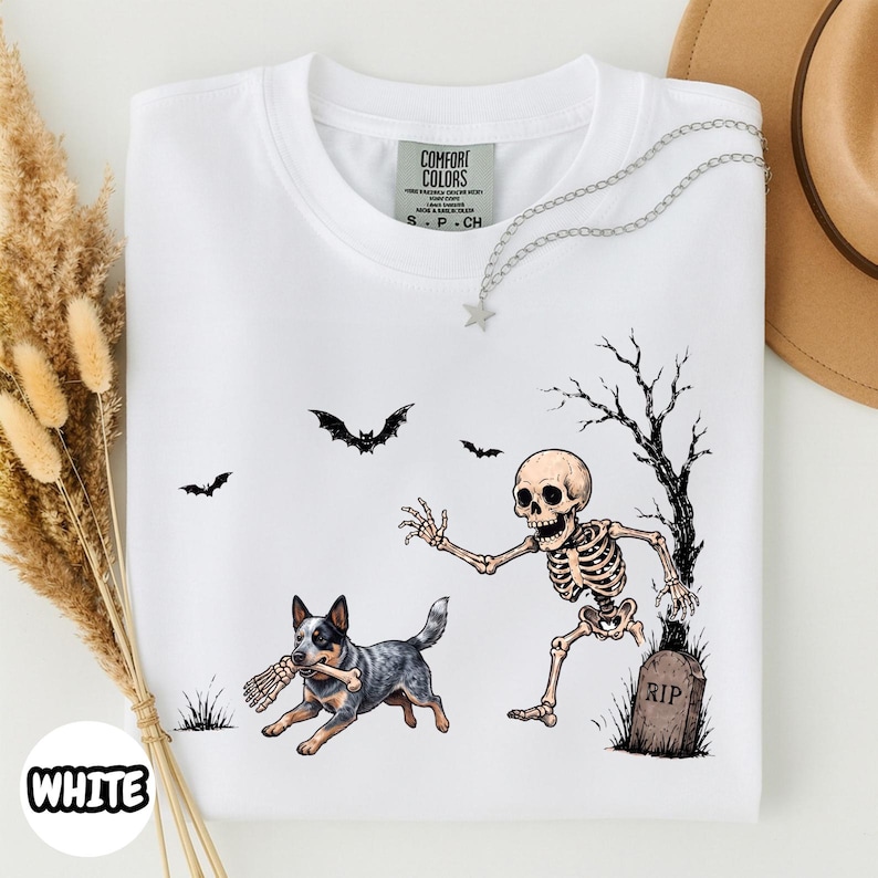 May include: A white t-shirt featuring a graphic print of a skeleton emerging from a grave near a bare tree, with a blue heeler dog running while holding a bone in its mouth. Two bats are flying in the background above the skeleton and dog.