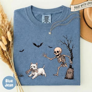 May include: A blue t-shirt featuring a graphic of a white West Highland Terrier dog holding a bone in its mouth, running away from a skeleton near a tombstone marked RIP, with two black bats flying in the background and a star-shaped necklace.