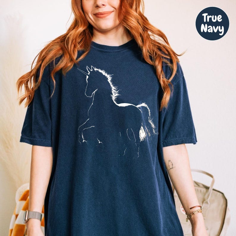 A woman with long red hair wearing a navy blue t-shirt with a white unicorn graphic.