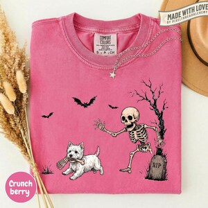 May include: A bright pink crew neck t-shirt featuring a graphic of a white dog holding a bone in its mouth, a skeleton emerging from a grave marked RIP, and black silhouettes of bats and a bare tree against the solid pink fabric background.