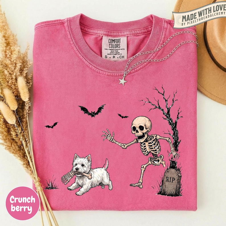 May include: A bright pink crew neck t-shirt featuring a graphic of a white dog holding a bone in its mouth, a skeleton emerging from a grave marked RIP, and black silhouettes of bats and a bare tree against the solid pink fabric background.