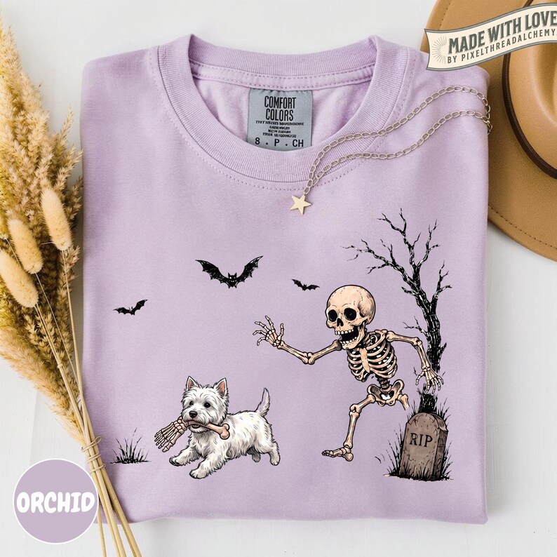May include: A light purple crew neck t-shirt featuring a graphic of a white West Highland Terrier dog holding a bone, a skeleton emerging from a grave marked RIP, and three black bats flying in the air. The shirt has a Comfort Colors brand label.