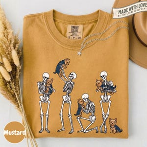 May include: A mustard yellow t-shirt featuring a graphic of four white skeletons interacting with small brown and black Yorkshire Terrier dogs. The shirt has a crew neckline and a Comfort Colors brand label printed on the inside of the collar.