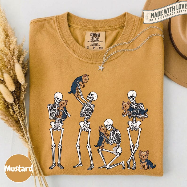 May include: A mustard yellow t-shirt featuring a graphic of four white skeletons interacting with small brown and black Yorkshire Terrier dogs. The shirt has a crew neckline and a Comfort Colors brand label printed on the inside of the collar.