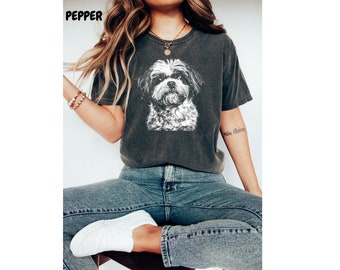 May include: Dark gray t-shirt featuring a detailed black and white illustration of a Shih Tzu dog. The word "PEPPER" is printed at the top left. The shirt is worn with blue jeans and white sneakers.