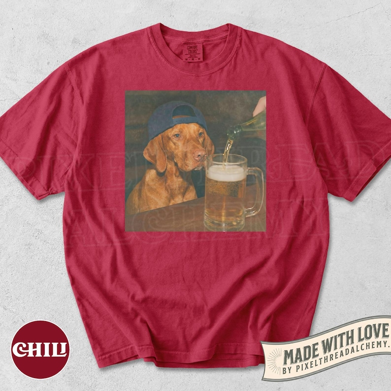 K&ouml;nnte beinhalten: Ein rotes Kurzarm-T-Shirt mit einem zentralen Grafikdruck eines braunen Hundes, der eine blaue Baseballkappe tr&auml;gt und zusieht, wie eine Fl&uuml;ssigkeit in einen gl&auml;sernen Bierkrug gegossen wird. Das T-Shirt hat einen Rundhalsausschnitt und eine Standardpassform.