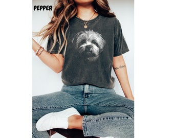 May include: A dark gray t-shirt with a white image of a dog's face on the front. The dog has a fluffy white coat and a black nose. The shirt is worn by a person with long blonde hair and a gold necklace.
