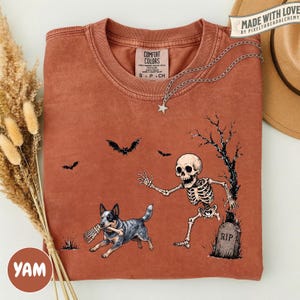 May include: A rust-colored t-shirt featuring a graphic of a skeleton reaching toward a blue heeler dog holding a bone in its mouth. The design includes a tombstone marked RIP, a bare tree, and two bats. The shirt has a Comfort Colors brand label.