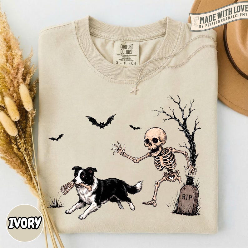 May include: A beige crewneck t-shirt featuring a graphic print of a black and white Border Collie dog holding a bone, running away from a human skeleton standing near a tombstone marked RIP, with bats and a bare tree in the background.