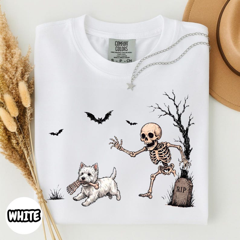 May include: A white t-shirt featuring a graphic of a skeleton emerging from a tombstone marked RIP, reaching toward a white West Highland Terrier dog holding a bone in its mouth, with two black bats flying in the background.