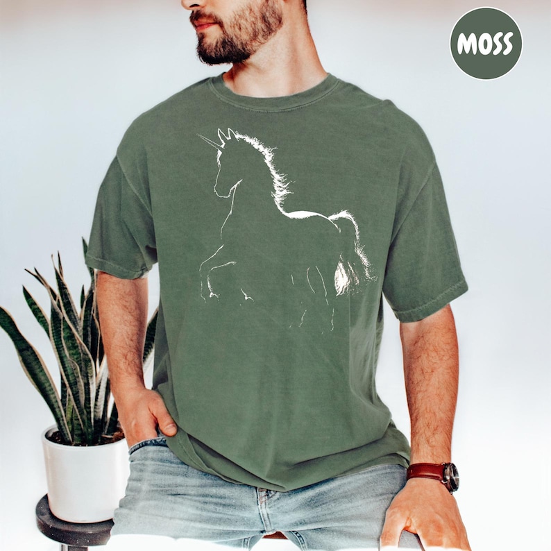 A man wearing a green t-shirt with a white graphic of a unicorn is sitting next to a potted plant.