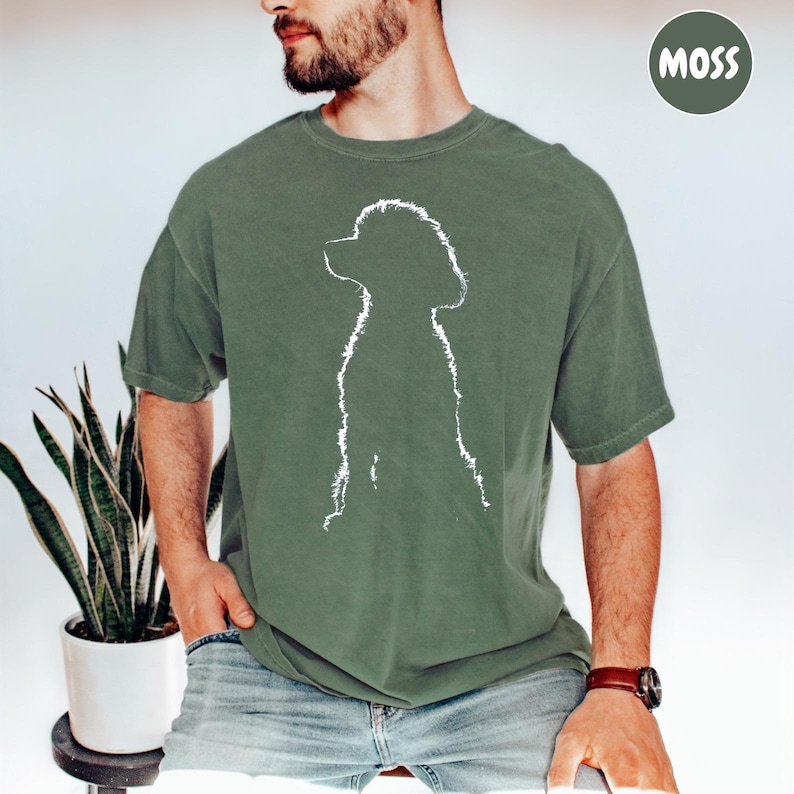 A man wearing a green t-shirt with a white silhouette of a dog is sitting in front of a potted plant.