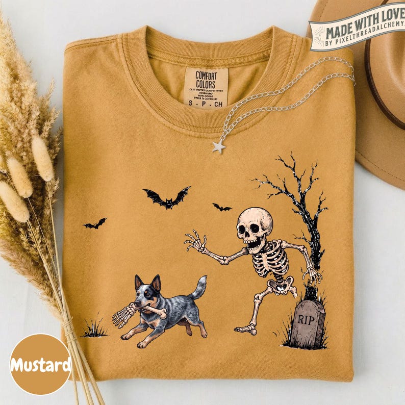 May include: A mustard yellow crew neck t-shirt featuring a graphic print of a skeleton reaching toward a blue heeler dog holding a bone in its mouth. The design includes a tombstone marked RIP, a bare tree, and two bats flying in the background.