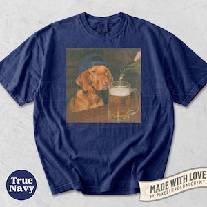 K&ouml;nnte beinhalten: Ein marineblaues T-Shirt mit einem Grafikdruck eines braunen Hundes, der eine dunkle Baseballkappe tr&auml;gt und neben einem Glaskrug sitzt, in den Bier aus einer Flasche gegossen wird. Das T-Shirt hat einen Rundhalsausschnitt und kurze &Auml;rmel.