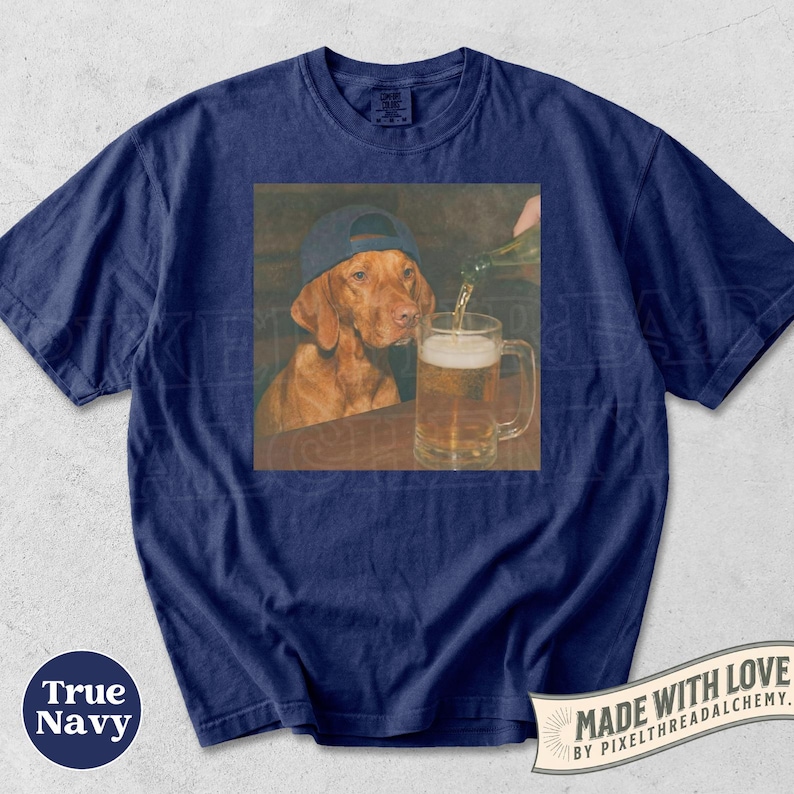 K&ouml;nnte beinhalten: Ein marineblaues T-Shirt mit einem Grafikdruck eines braunen Hundes, der eine dunkle Baseballkappe tr&auml;gt und neben einem Glaskrug sitzt, in den Bier aus einer Flasche gegossen wird. Das T-Shirt hat einen Rundhalsausschnitt und kurze &Auml;rmel.
