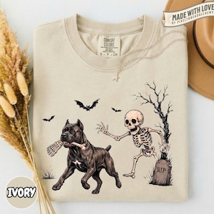 May include: A beige crew neck t-shirt featuring a graphic print of a skeleton running towards a dark-coloured dog that is holding a bone in its mouth. The design includes a bare tree, a tombstone labelled RIP, and two flying bats in black ink.