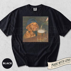 K&ouml;nnte beinhalten: Ein schwarzes Kurzarm-T-Shirt mit einem Grafikdruck eines braunen Hundes, der eine blaue Baseballkappe tr&auml;gt und neben einem mit Bier gef&uuml;llten Glaskrug sitzt. Das T-Shirt hat einen Rundhalsausschnitt und ein schwarzes Farbetikett.