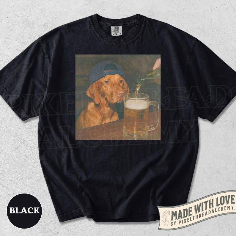 K&ouml;nnte beinhalten: Ein schwarzes Kurzarm-T-Shirt mit einem Grafikdruck eines braunen Hundes, der eine blaue Baseballkappe tr&auml;gt und neben einem mit Bier gef&uuml;llten Glaskrug sitzt. Das T-Shirt hat einen Rundhalsausschnitt und ein schwarzes Farbetikett.