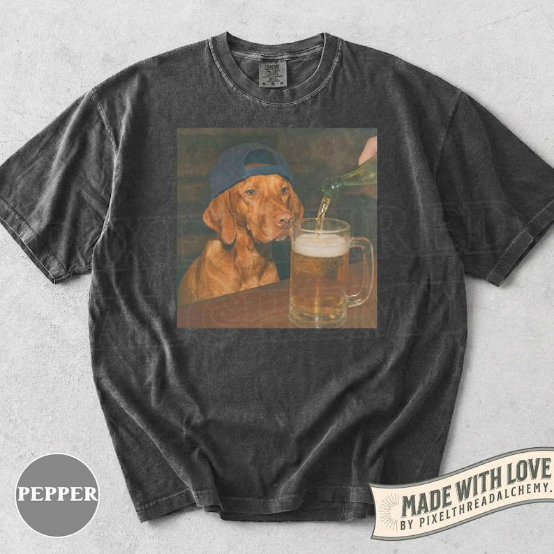K&ouml;nnte beinhalten: Ein anthrazitfarbenes Kurzarm-T-Shirt mit einem Grafikdruck eines braunen Hundes, der eine blaue Baseballkappe tr&auml;gt und zusieht, wie eine klare Fl&uuml;ssigkeit in einen Bierkrug gegossen wird. Das Shirt hat einen Rundhalsausschnitt und Stoffoptik.