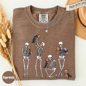 May include: A brown crewneck t-shirt featuring a graphic of four human skeletons interacting with small brown and black Yorkshire Terrier dogs. The shirt has a Comfort Colors label at the inner neckline and a silver star-shaped pendant necklace.