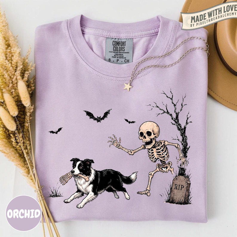 May include: A light purple t-shirt featuring a graphic of a black and white dog holding a bone in its mouth while running from a skeleton near a tombstone marked RIP. The design includes two bats and a bare tree. The shirt has a crew neck collar.