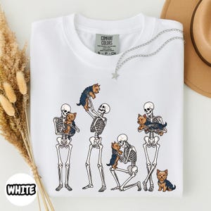 May include: A white t-shirt featuring a graphic print of four human skeletons interacting with small brown and black Yorkshire Terrier dogs. The skeletons are shown standing, kneeling, and holding the dogs. The shirt has a Comfort Colors label.
