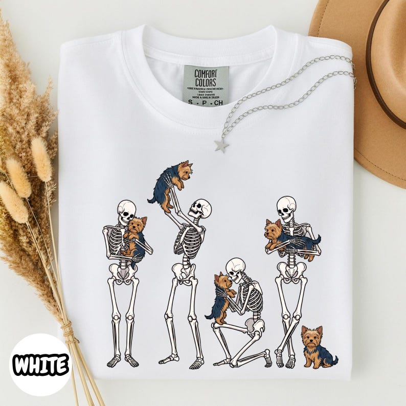 May include: A white t-shirt featuring a graphic print of four human skeletons interacting with small brown and black Yorkshire Terrier dogs. The skeletons are shown standing, kneeling, and holding the dogs. The shirt has a Comfort Colors label.