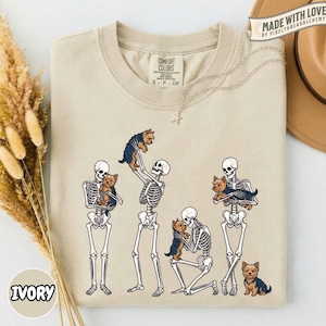 May include: An ivory crew neck t-shirt featuring a graphic print of four human skeletons interacting with small brown and black Yorkshire Terrier dogs. The skeletons are shown standing, kneeling, and holding the dogs in various poses.