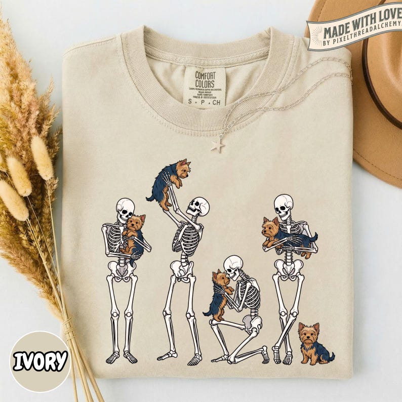 May include: An ivory crew neck t-shirt featuring a graphic print of four human skeletons interacting with small brown and black Yorkshire Terrier dogs. The skeletons are shown standing, kneeling, and holding the dogs in various poses.