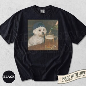 May include: Black t-shirt featuring a square image of a white dog wearing a blue baseball cap, sitting at a table while beer is poured into a mug. The shirt has the word "BLACK" printed on the bottom left.