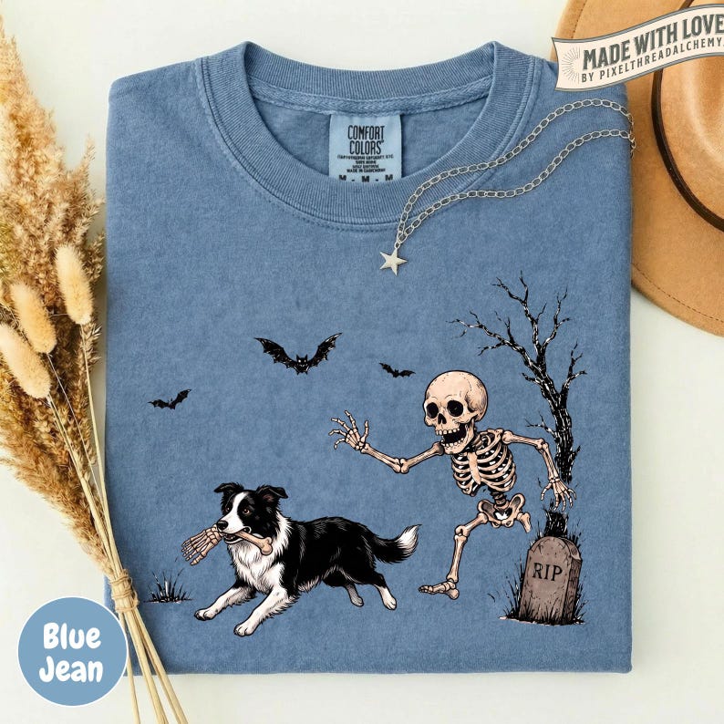 May include: A blue t-shirt featuring a graphic of a black and white dog holding a bone in its mouth, running from a skeleton near a tombstone marked RIP. The design includes two bats and a bare tree. A silver star necklace rests on the shirt.