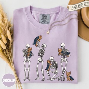 May include: A light purple crew neck t-shirt featuring a graphic print of four white skeletons interacting with small brown and black Yorkshire Terrier dogs. The shirt has a Comfort Colors brand label at the inner neckline and a silver star necklace.