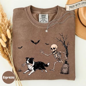 May include: A brown crewneck t-shirt featuring a graphic of a black and white dog holding a bone in its mouth, a skeleton reaching toward the dog, a tombstone marked RIP, and bats flying in the air. The shirt has a Comfort Colors brand label.