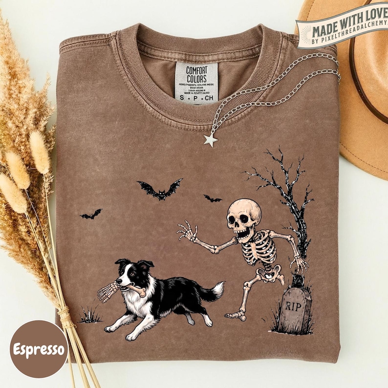 May include: A brown crewneck t-shirt featuring a graphic of a black and white dog holding a bone in its mouth, a skeleton reaching toward the dog, a tombstone marked RIP, and bats flying in the air. The shirt has a Comfort Colors brand label.