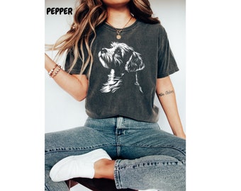 May include: A dark gray t-shirt with a white silhouette of a dog's head. The dog is looking up and to the right. The shirt is being worn by a person with long blonde hair. The person is sitting down and wearing blue jeans.