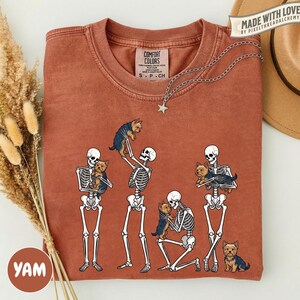 May include: A rust-colored crew neck t-shirt featuring a graphic print of four white skeletons interacting with small brown Yorkshire Terrier dogs. The shirt has a Comfort Colors brand label at the inner neckline and a short-sleeved design.