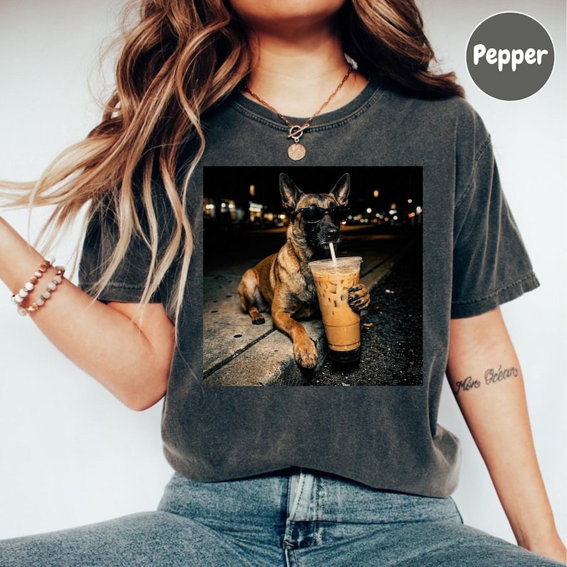 May include: Dark gray t-shirt featuring a square image of a dog wearing sunglasses and holding an iced coffee. The dog is a Belgian Malinois, and the image is set against a blurred night scene.