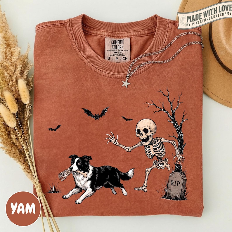 May include: A rust-colored short-sleeve t-shirt featuring a graphic of a skeleton reaching toward a black and white border collie holding a bone in its mouth. The design includes a tombstone marked RIP, a bare tree, and two flying bats.