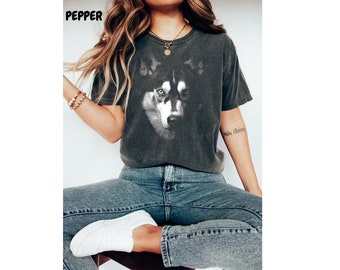 May include: A woman wearing a dark grey t-shirt with a black and white husky dog graphic. The shirt has a crew neck and short sleeves. The woman is wearing blue jeans and white sneakers.