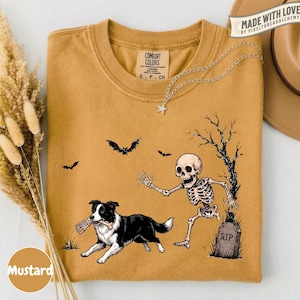 May include: A mustard yellow t-shirt featuring a graphic of a skeleton reaching toward a black and white Border Collie dog holding a bone in its mouth. The design includes a tombstone marked RIP, a bare tree, and two flying bats. The shirt has a Comfort Colors label.