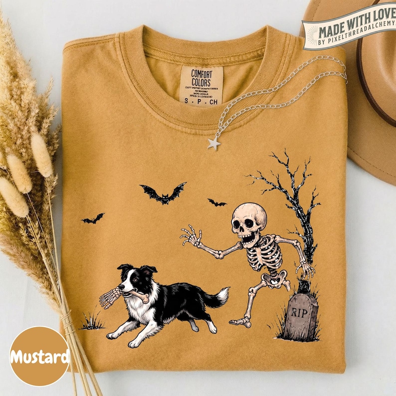 May include: A mustard yellow t-shirt featuring a graphic of a skeleton reaching toward a black and white Border Collie dog holding a bone in its mouth. The design includes a tombstone marked RIP, a bare tree, and two flying bats. The shirt has a Comfort Colors label.