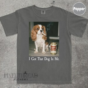 A t-shirt with a graphic design featuring a brown and white dog sitting on a wooden surface, smoking a cigarette, and next to a can of Miller High Life beer.