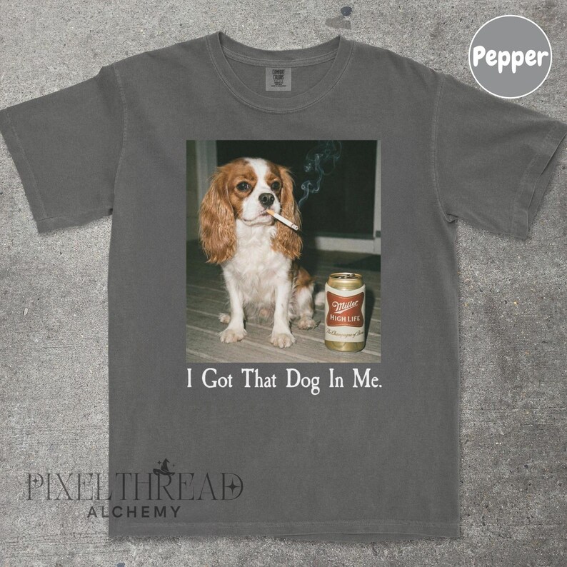 A t-shirt with a graphic design featuring a brown and white dog sitting on a wooden surface, smoking a cigarette, and next to a can of Miller High Life beer.