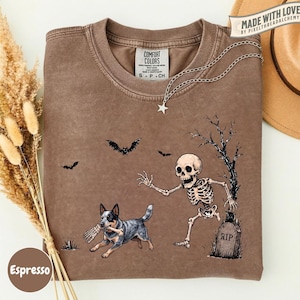 May include: A brown short-sleeve t-shirt featuring a graphic print of a skeleton running toward a tombstone marked RIP, while a dog carries a bone in its mouth. Bats fly in the background near a bare tree. The shirt has a Comfort Colors label.