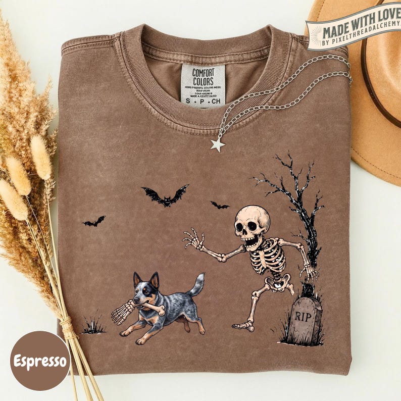 May include: A brown short-sleeve t-shirt featuring a graphic print of a skeleton running toward a tombstone marked RIP, while a dog carries a bone in its mouth. Bats fly in the background near a bare tree. The shirt has a Comfort Colors label.
