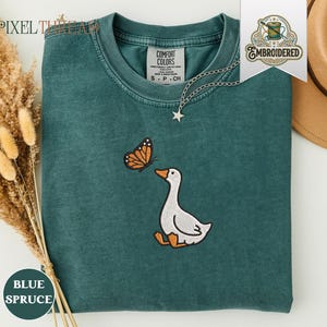 Puede incluir: Una sudadera de cuello redondo color azul abeto con un diseño bordado de un ganso blanco con patas y pico naranja mirando hacia una mariposa naranja y negra. La prenda tiene un cuello acanalado y una etiqueta de la marca Comfort Colors en el escote.