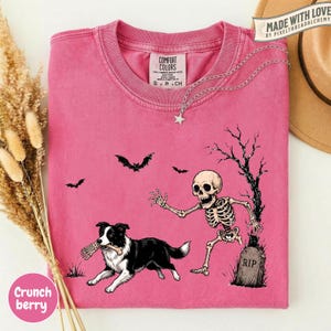 May include: A pink short-sleeve t-shirt featuring a graphic of a skeleton chasing a black and white Border Collie dog holding a bone. The design includes a tombstone labeled RIP, a bare tree, and two bats. The shirt has a Comfort Colors brand label.