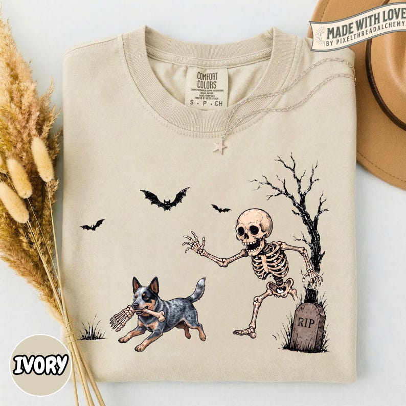 May include: A beige t-shirt featuring a graphic print of a skeleton running from a dog that is holding a bone in its mouth. The design includes a tombstone labeled RIP, a bare tree, and three flying bats. The shirt has a crew neckline and short sleeves.